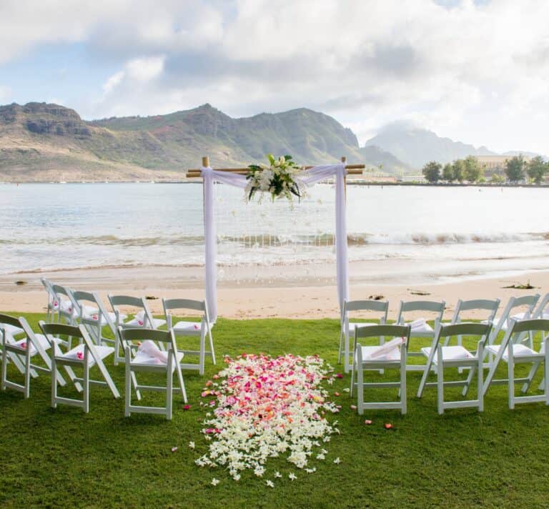 Beachfront wedding ceremony setup at The Royal Sonesta Kauai Resort, featuring white chairs arranged on green grass, a floral arch, and a scenic ocean and mountain backdrop.