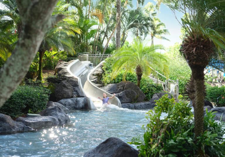 Tropical waterslide at The Royal Sonesta Kauai Resort, surrounded by lush greenery and palm trees, with a guest splashing into the pool below.