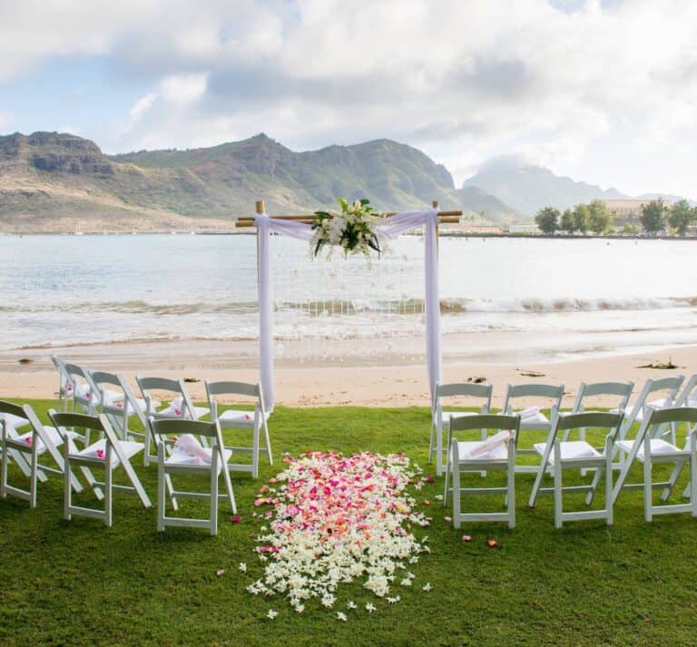 Beachfront wedding ceremony setup at The Royal Sonesta Kauai Resort, featuring white chairs arranged on green grass, a floral arch, and a scenic ocean and mountain backdrop.