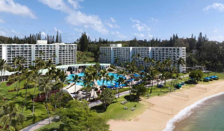 Aerial view of The Royal Sonesta Kauai Resort with two large white buildings, palm trees, swimming pools, lounge chairs, and a sandy beach under a clear sky.